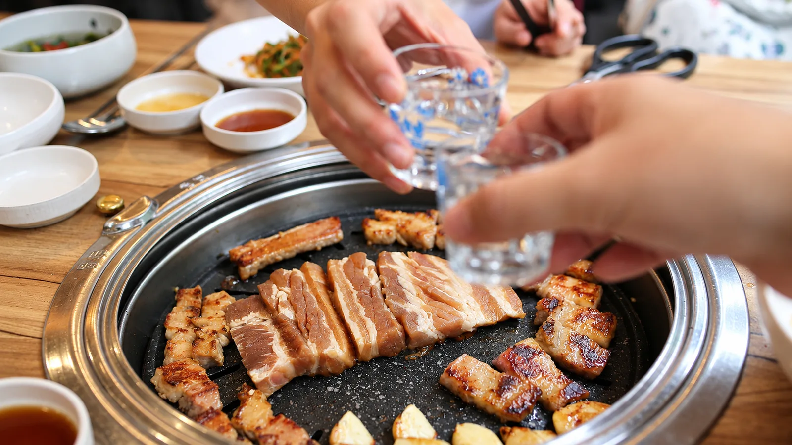 불판 위 돼지갈비를 앞에 두고 소주잔을 부딪치는 손, 곁들임 반찬